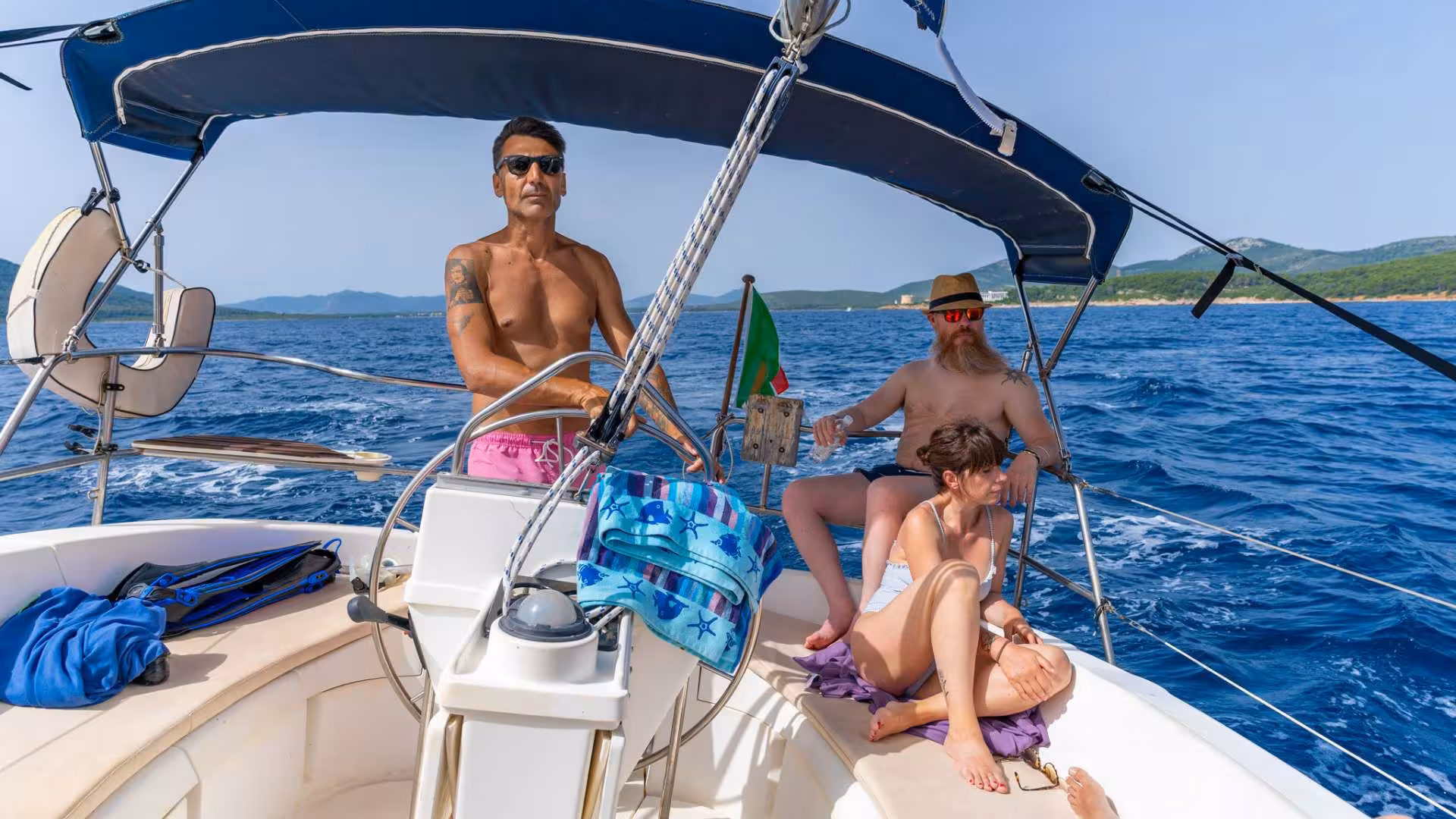 Captain steering a sailboat with passengers enjoying the scenic views of Porto Conte Park, Alghero.