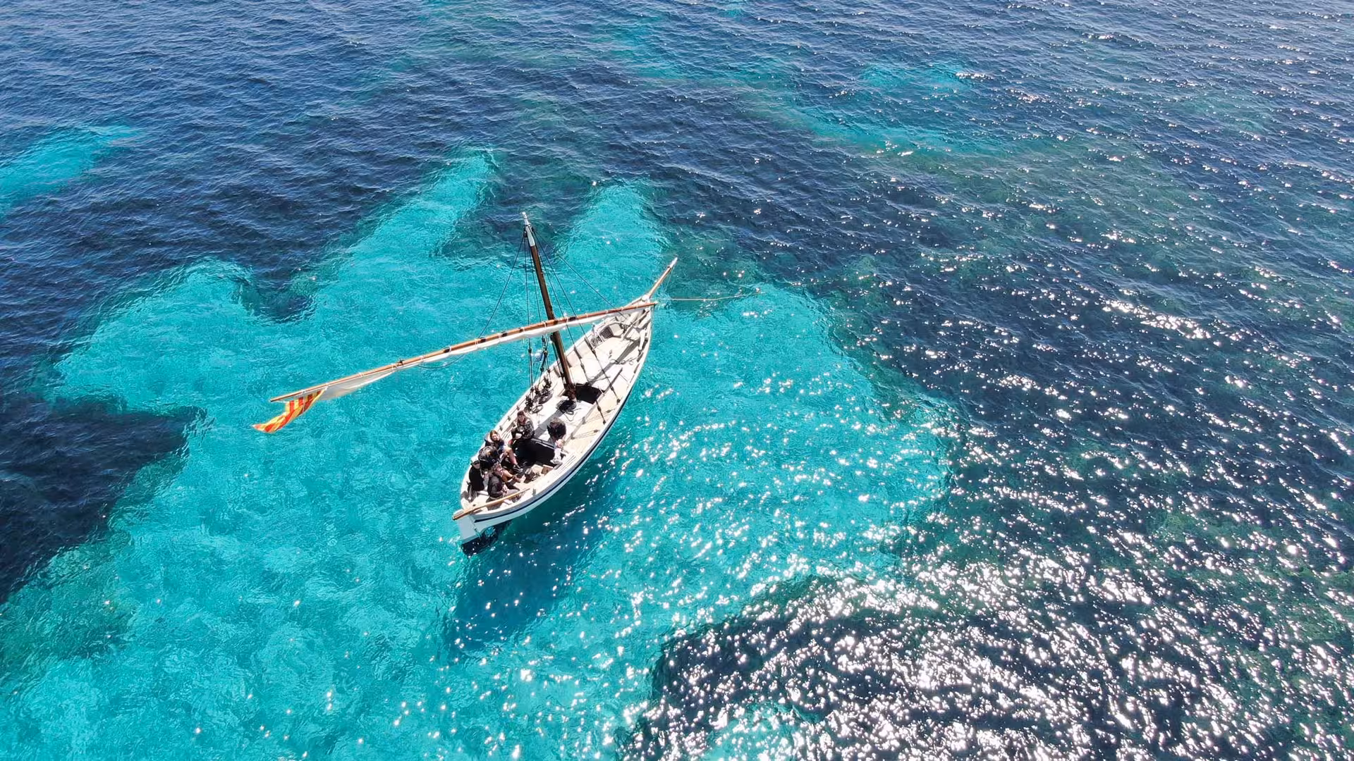 Aerial view of a sailboat gliding over the vibrant turquoise waters off the coast of Alghero during a scenic tour.