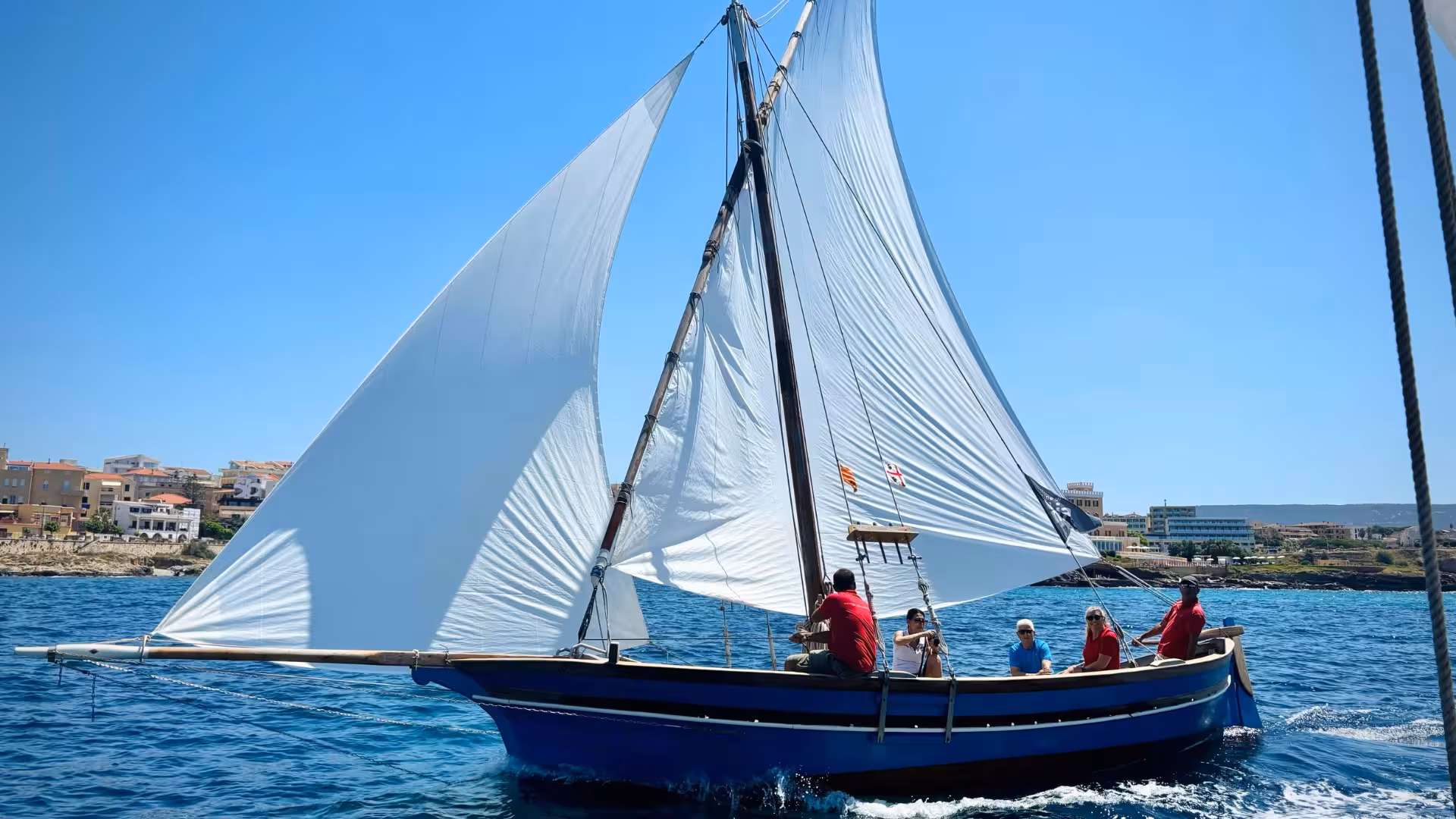 Sailboat with white sails cruising near Alghero's coastline on a sunny day, showcasing a scenic Sardinian sailing tour.