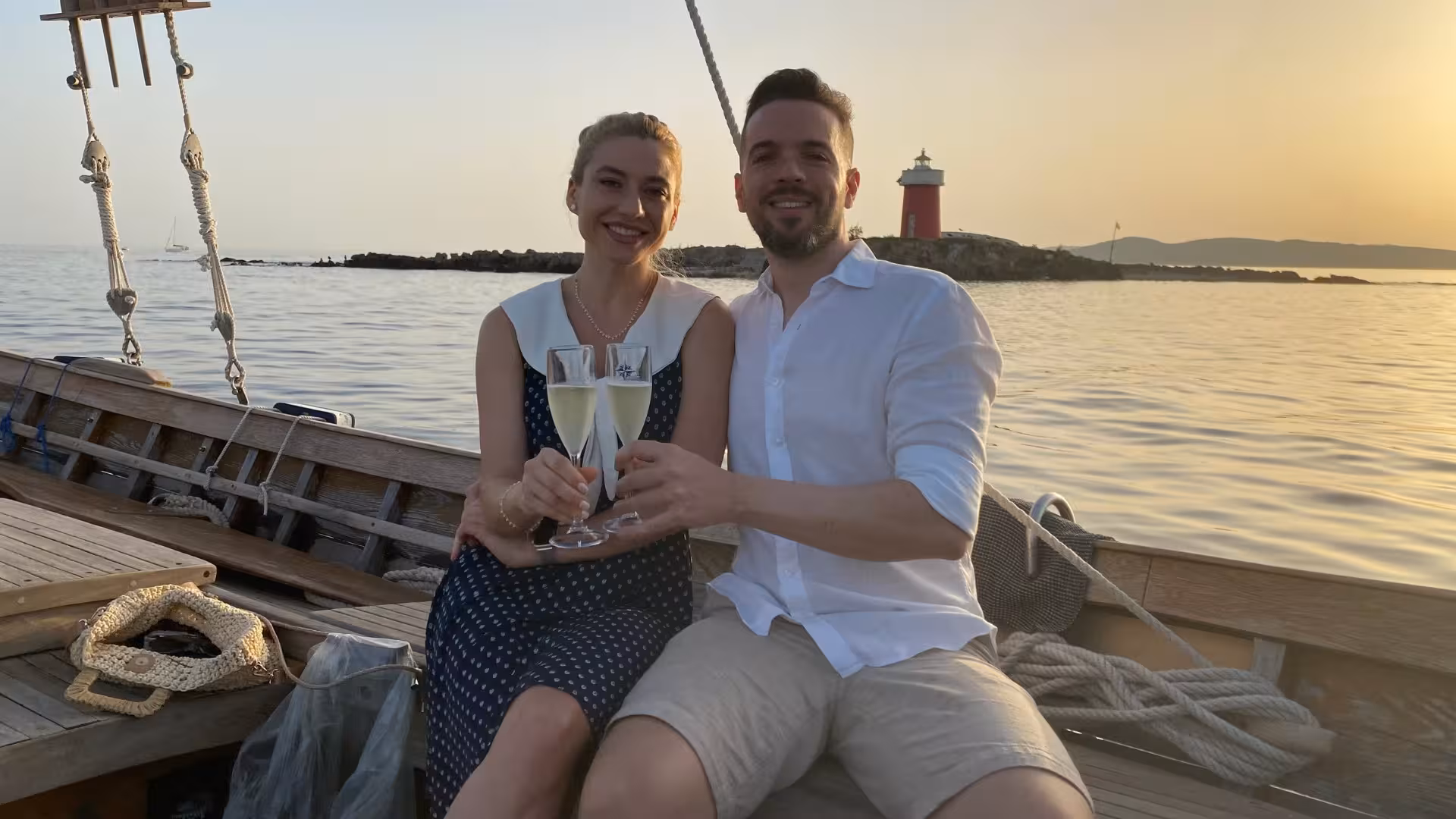 Couple toasting with champagne on a romantic sunset sailing tour in Alghero, near a lighthouse.