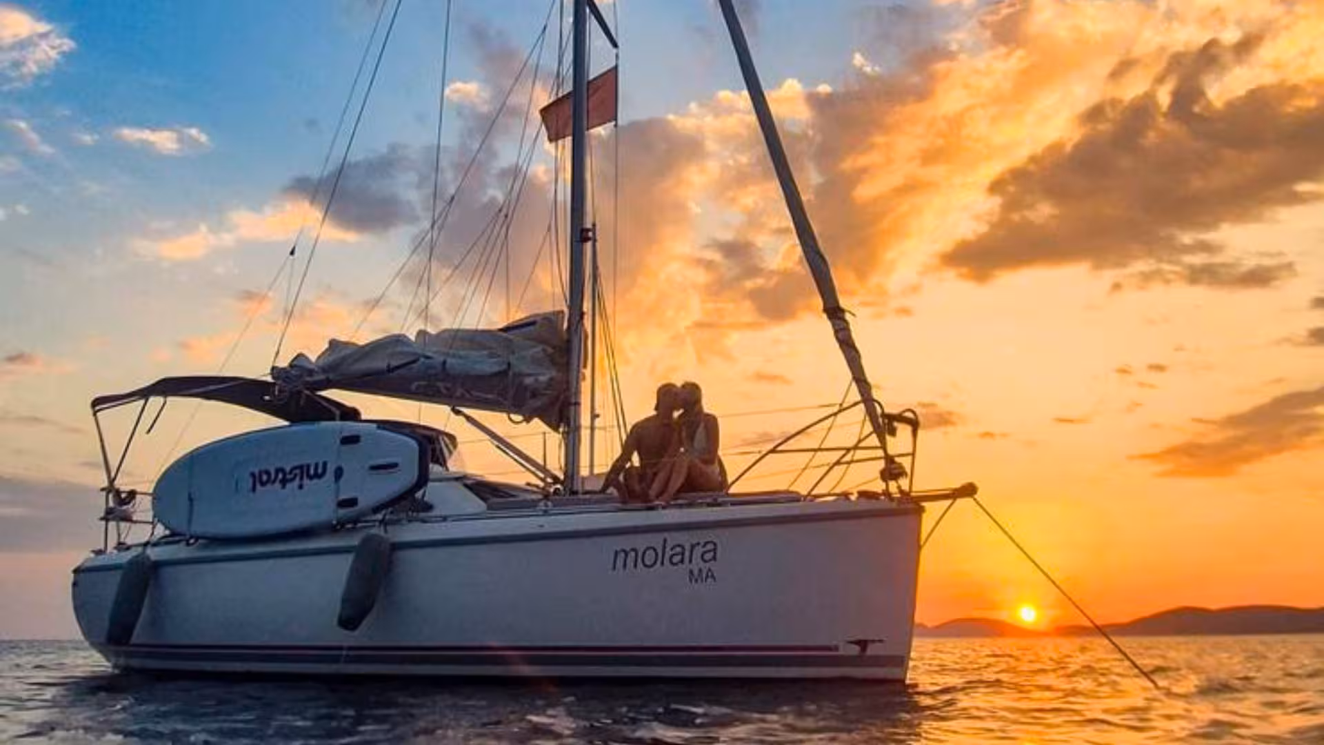 Couple sitting on a sailboat named Molara during a romantic Alghero sunset sailing tour, with colorful skies.