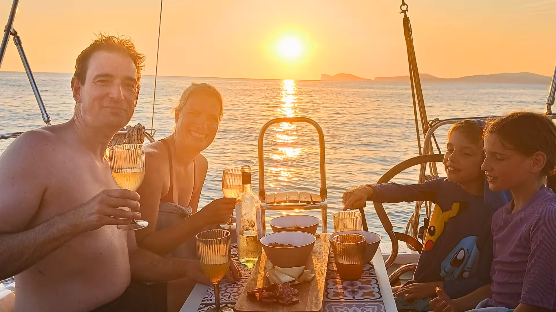 Family enjoying a sunset dinner on a sailing tour from Alghero, with glasses raised against a golden horizon.