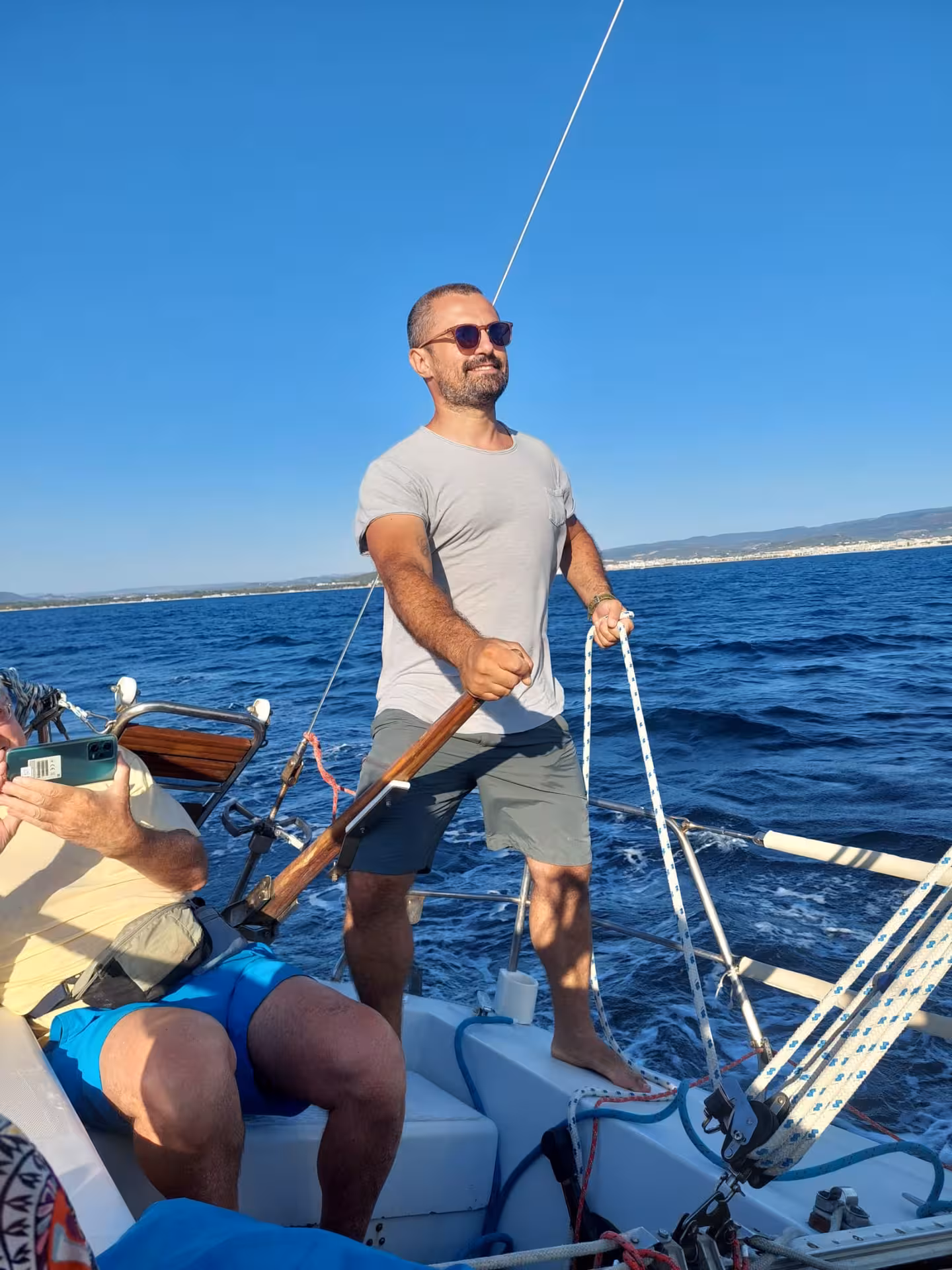 Man steering sailboat on Alghero sunset sailing tour from Fertilia under clear blue skies.