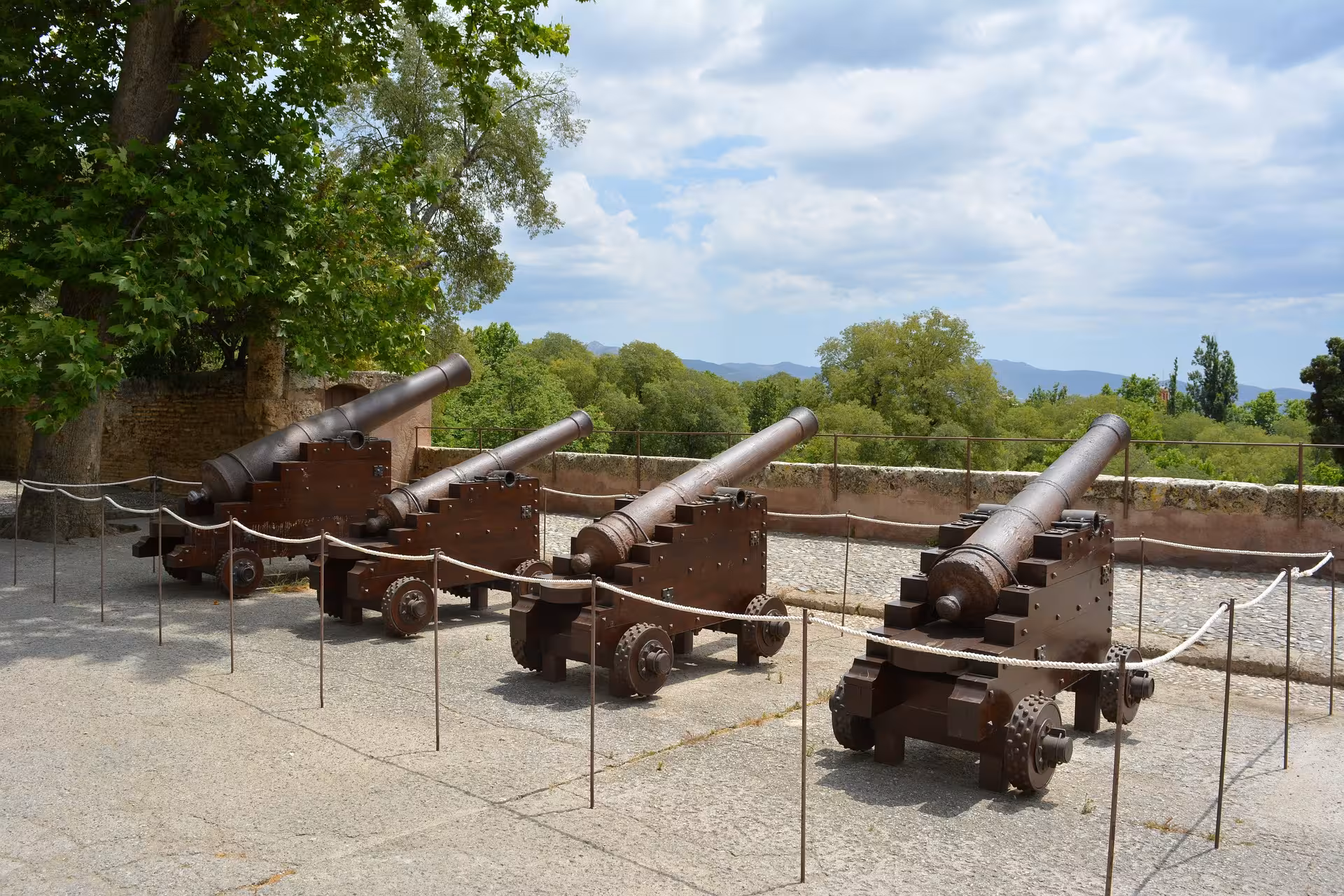 Row of historic cannons displayed on the grounds of the Alhambra, set against lush greenery and a clear sky.