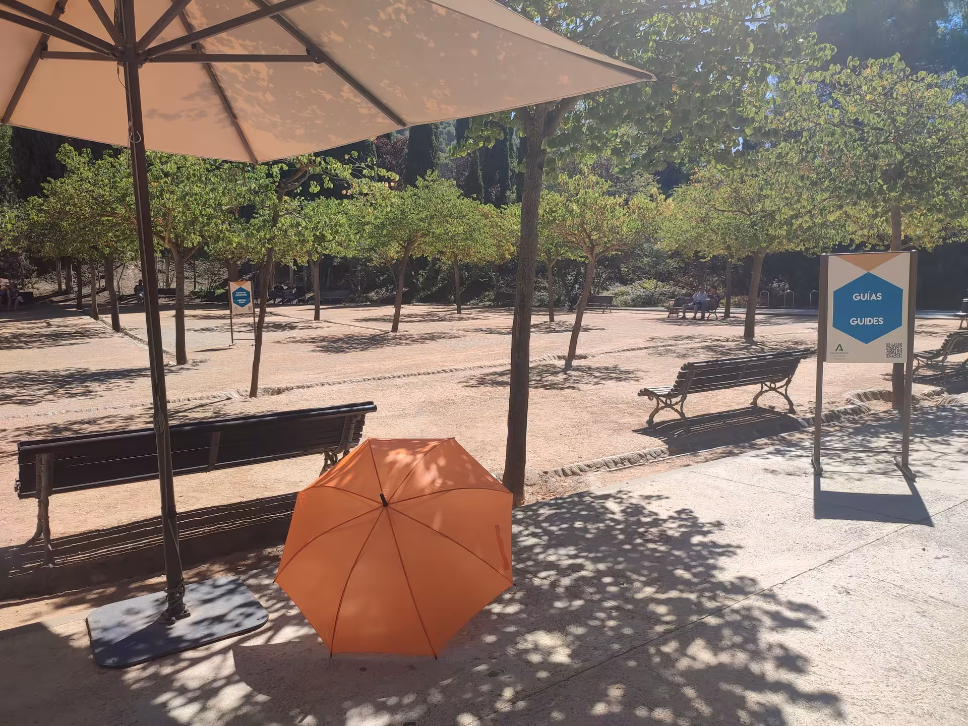 Guided tour meeting point at Alhambra with benches, orange umbrella, and signage under the shade of trees.
