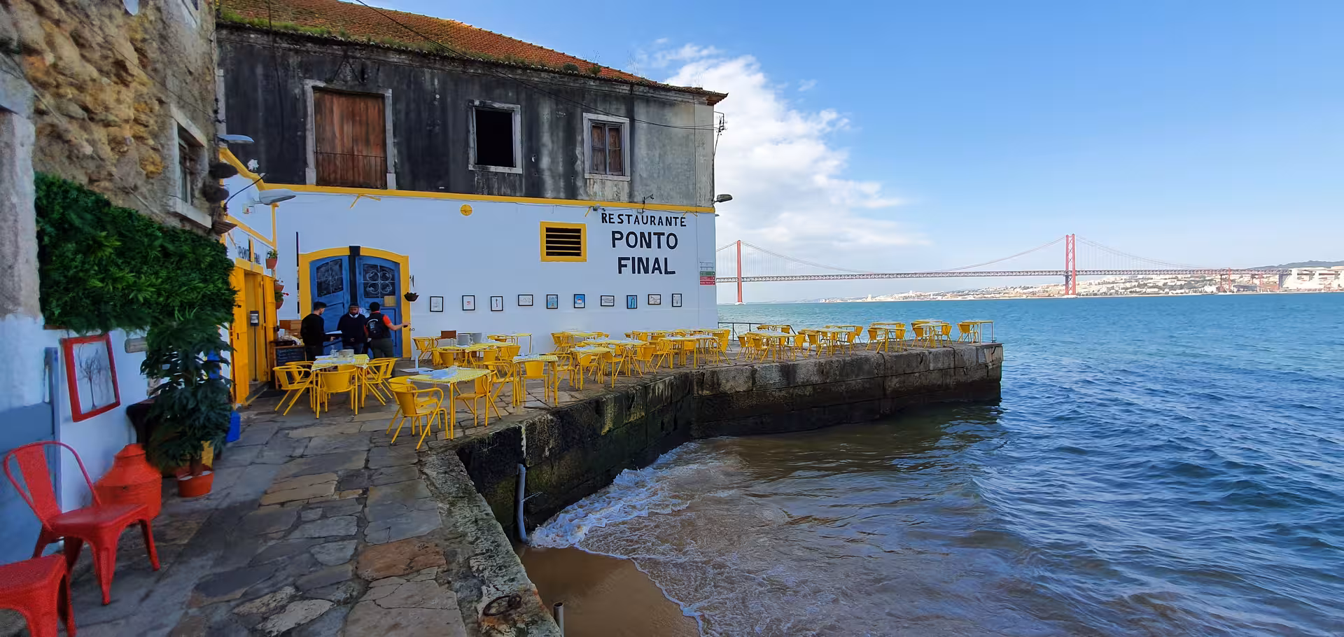 Charming waterfront restaurant in Almada with vibrant yellow chairs overlooking the Tagus River and 25 de Abril Bridge.