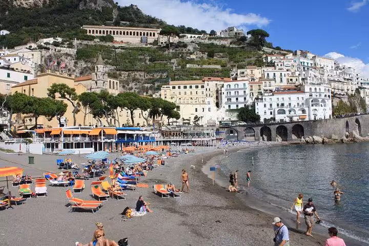 Scenic Amalfi beach view with colorful sun loungers, visitors enjoying the seaside, and picturesque hillside buildings.