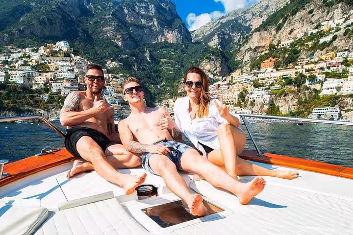 Group of friends relaxing on a boat with Positano's colorful cliffside buildings in the background on the Amalfi Coast.