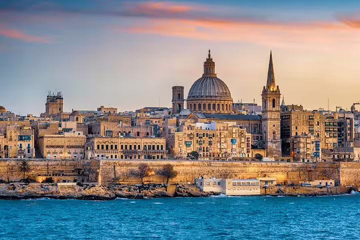 Coastal cityscape with domed cathedral and historic limestone buildings overlooking blue Mediterranean waters at sunset