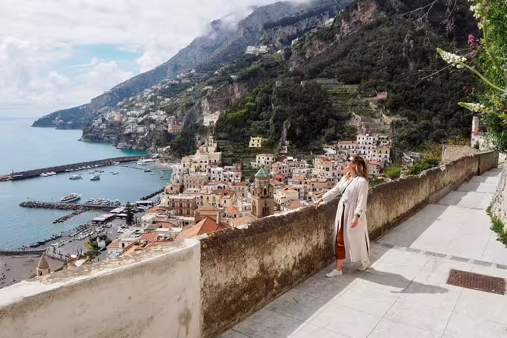 Woman overlooking Amalfi Coast town with colorful buildings, harbor, and green cliffs on a cloudy day