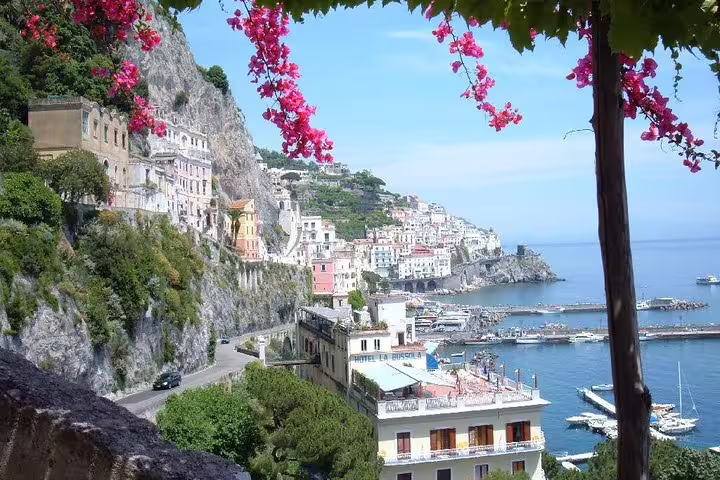 Scenic Amalfi Coast shore excursion view with cliffside pastel houses, marina, and Tyrrhenian Sea framed by pink bougainvillea
