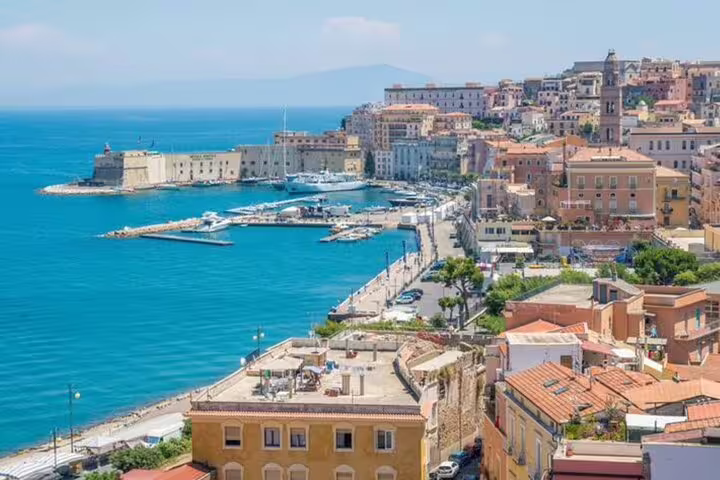 Aerial view of a sunny Italian coastal town harbor with colorful buildings, marina, and blue Mediterranean sea