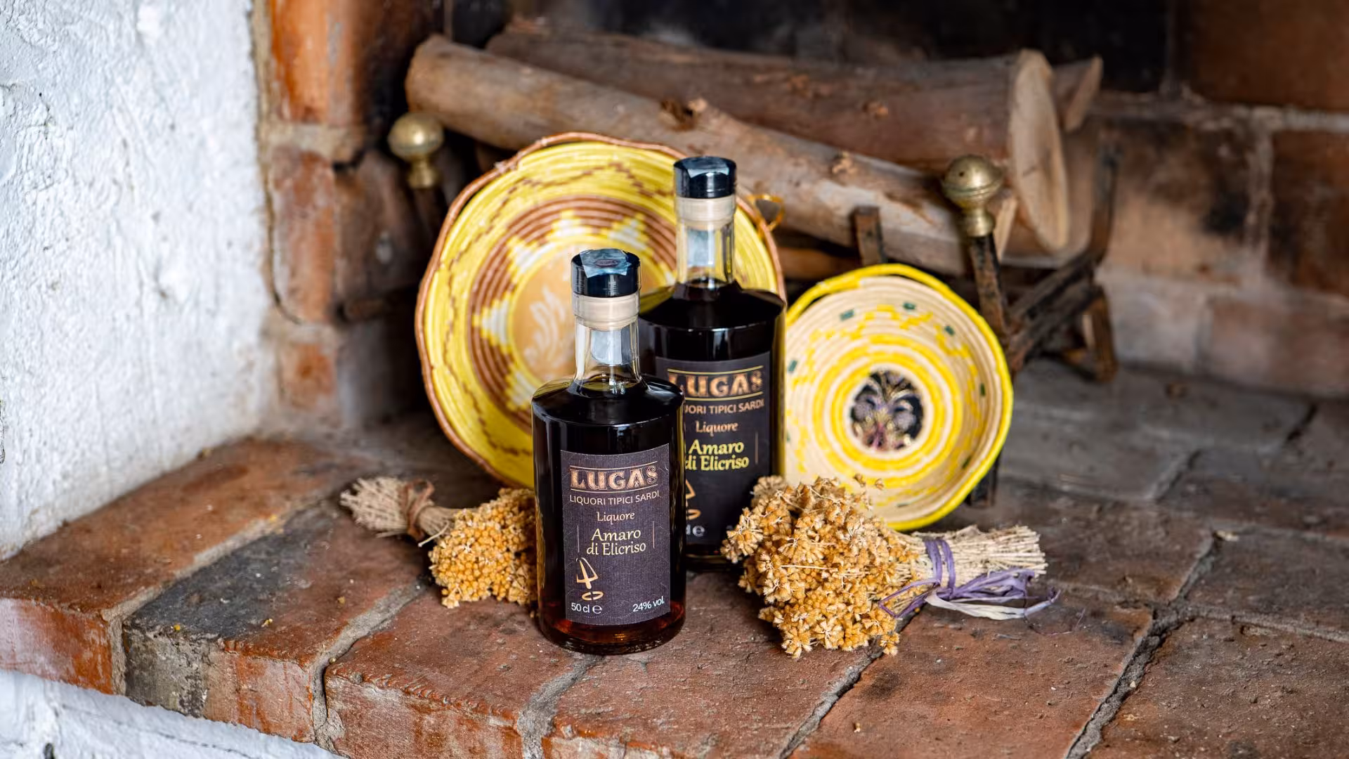 Two bottles of Amaro di Elicriso by Lugas in a rustic setting with decorative plates and dried flowers.