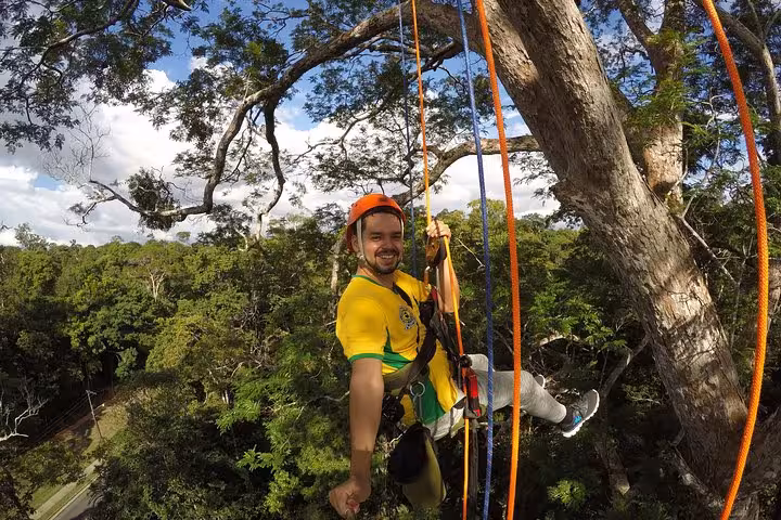 Smiling guide on ropes beside giant Amazon tree, showcasing canopy tree climbing and rainforest views
