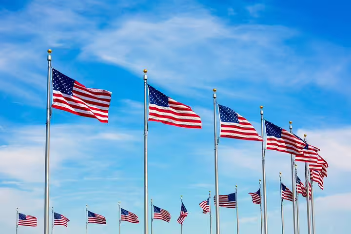 Multiple American flags on tall poles waving against a bright blue sky in Washington DC