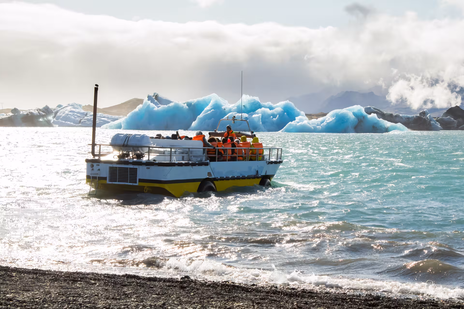 Amphibious tour boat with passengers in orange life jackets navigating Iceland's Glacier Lagoon among blue icebergs