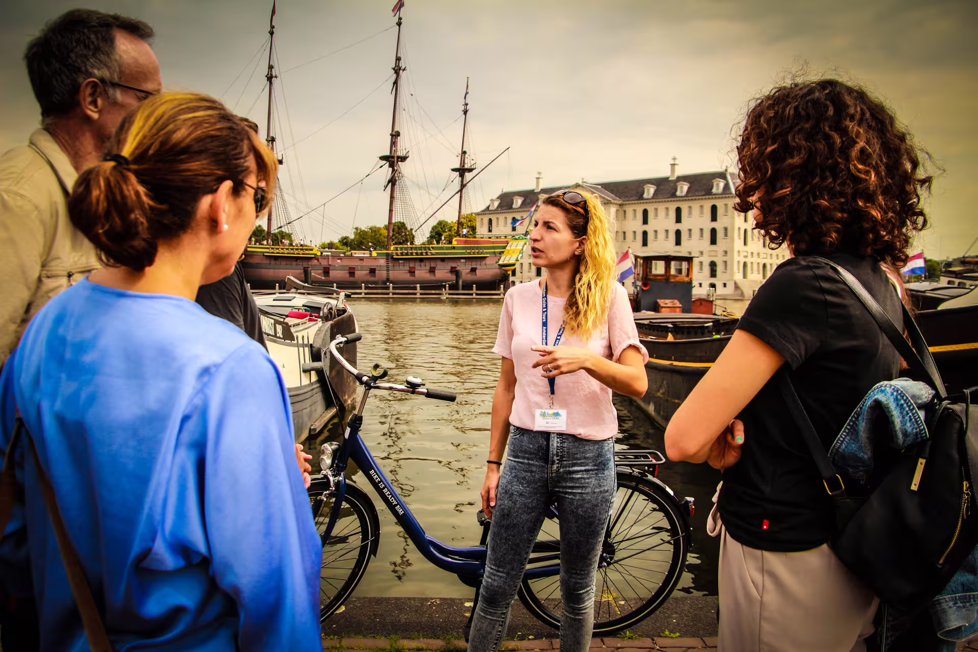 Tour group with guide and bicycles by Amsterdam canal, enjoying scenic views of historic ships and architecture.