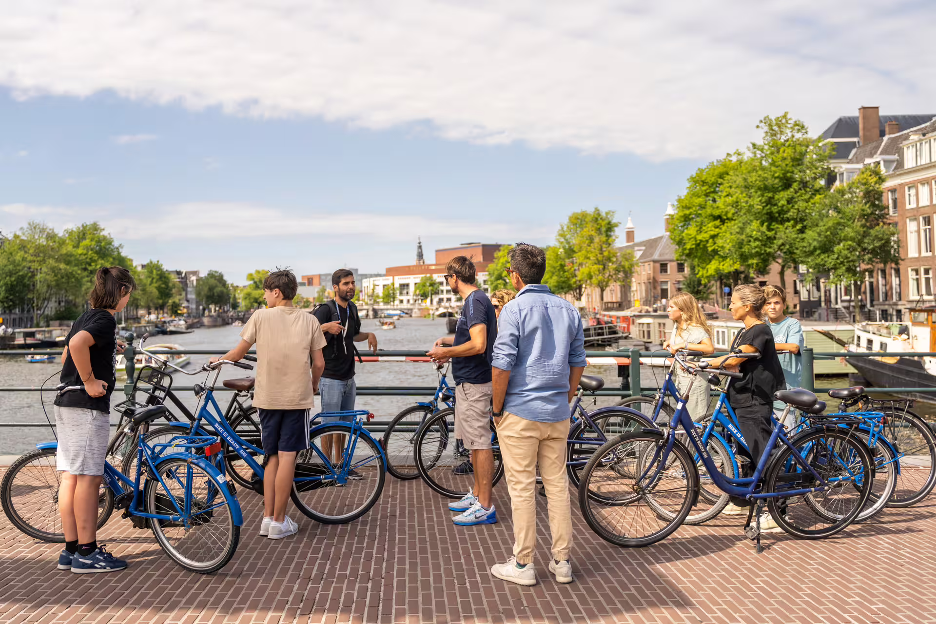 Group of tourists on blue bikes enjoying a sunny day along an Amsterdam canal, perfect for a tour with cheese and drinks.