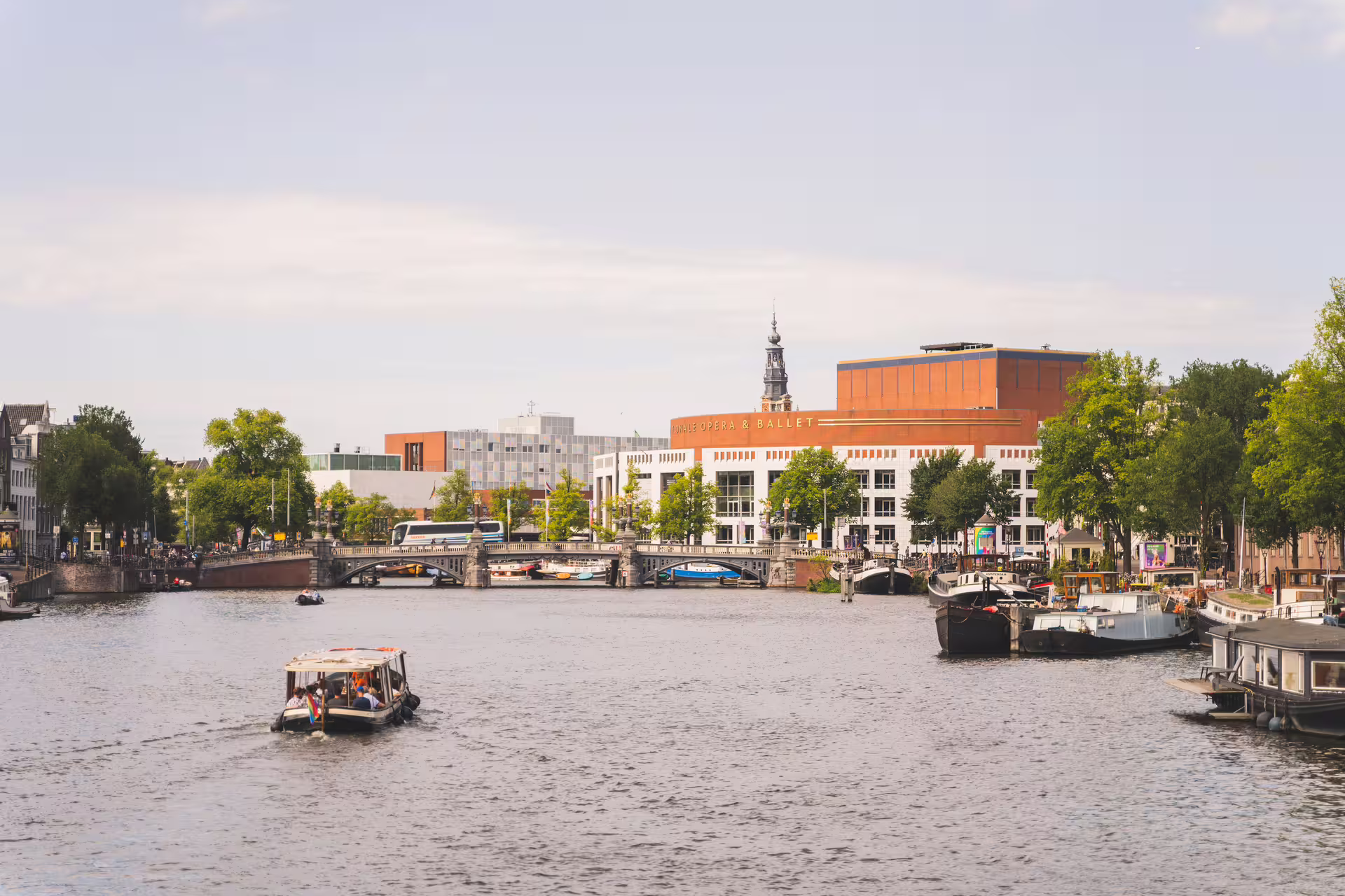 Scenic Amsterdam canal view featuring boats, Royal Opera House, and lush greenery, ideal for bike tour and canal cruise experience.