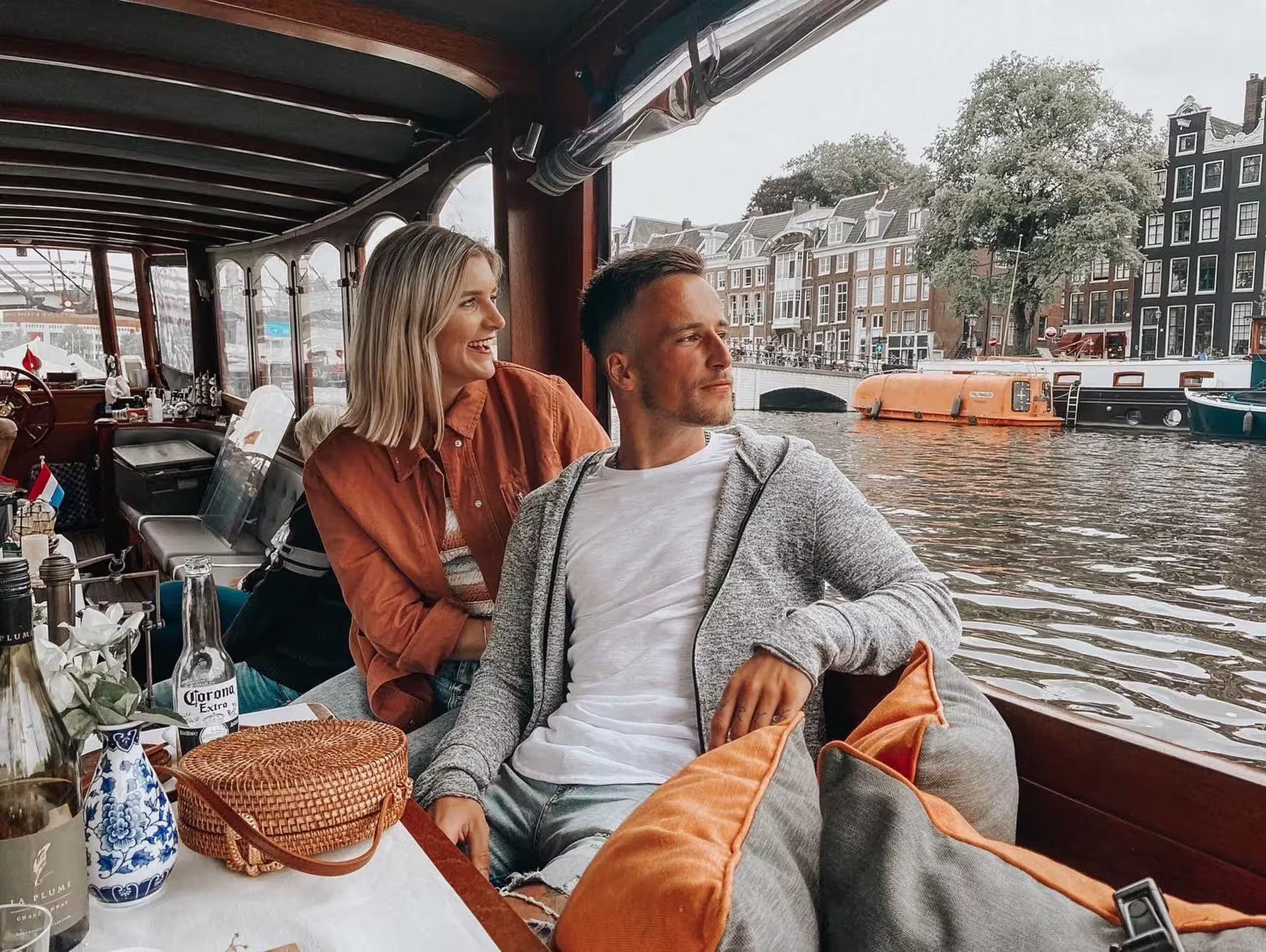 Couple enjoying Amsterdam canal cruise with drinks and cheese tasting, showcasing a scenic view of historic canals and architecture.