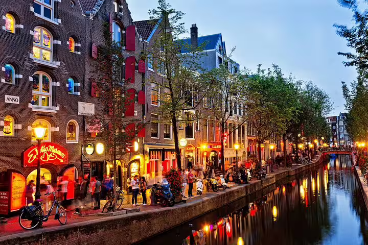 Amsterdam canal at dusk with illuminated historic buildings and people strolling along the Red Light District waterway