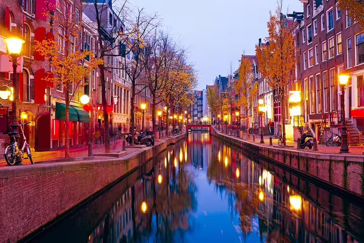 Amsterdam canal at dusk with glowing street lamps reflecting on water, lined with trees and historic brick buildings