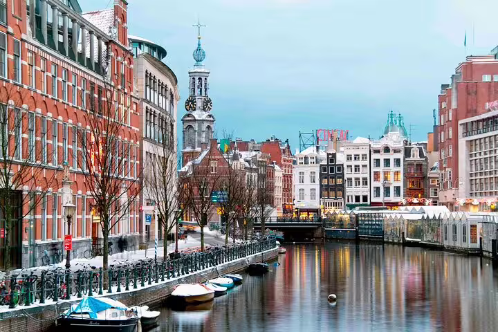 Amsterdam canal with snow-dusted buildings, moored boats, and Munttoren clock tower in winter