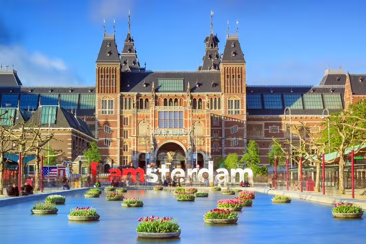 Rijksmuseum with I Amsterdam sign and colorful tulip planters in reflecting pool under blue sky