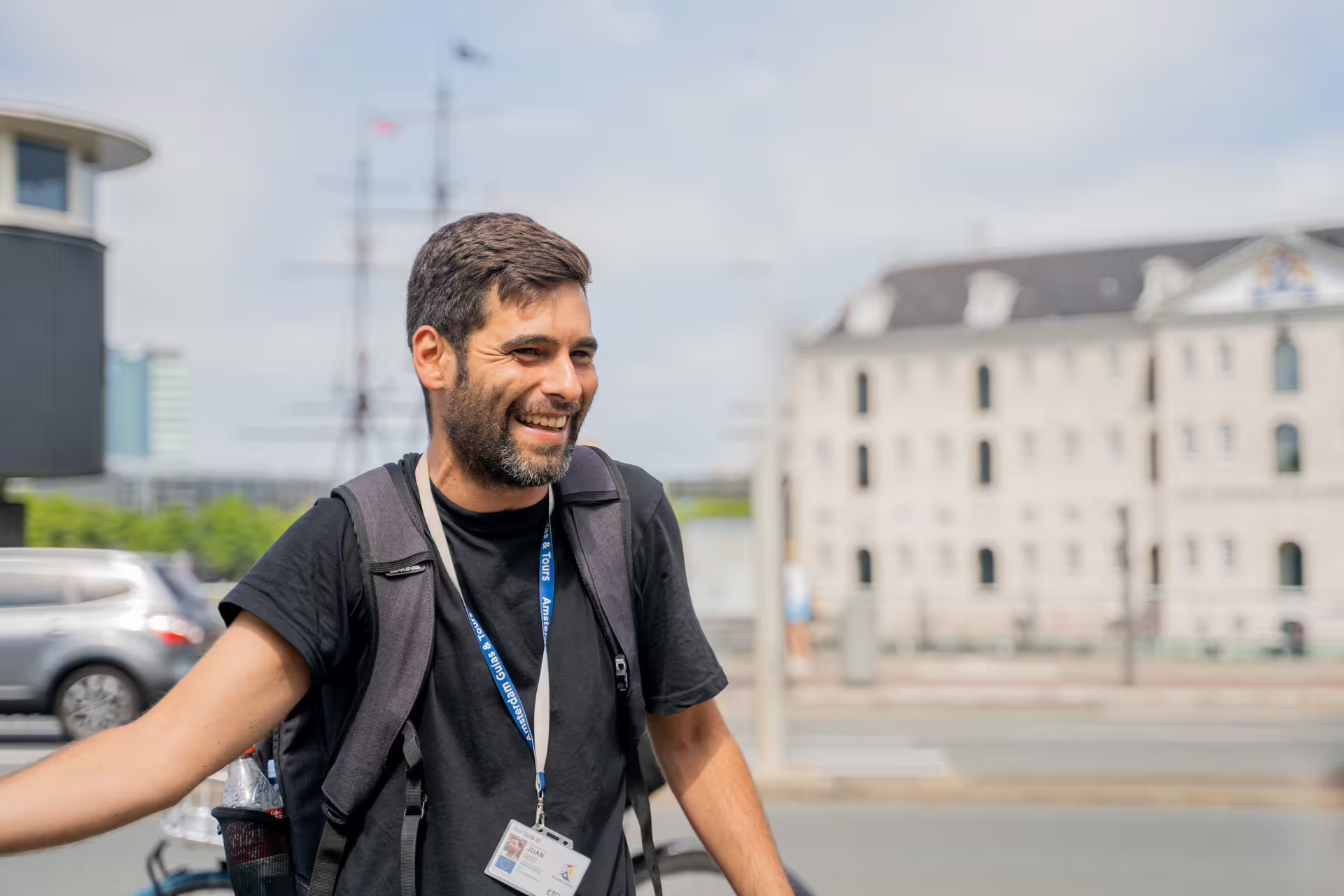 Smiling tour guide with a backpack leads a countryside private bike tour near historic buildings on a sunny day.