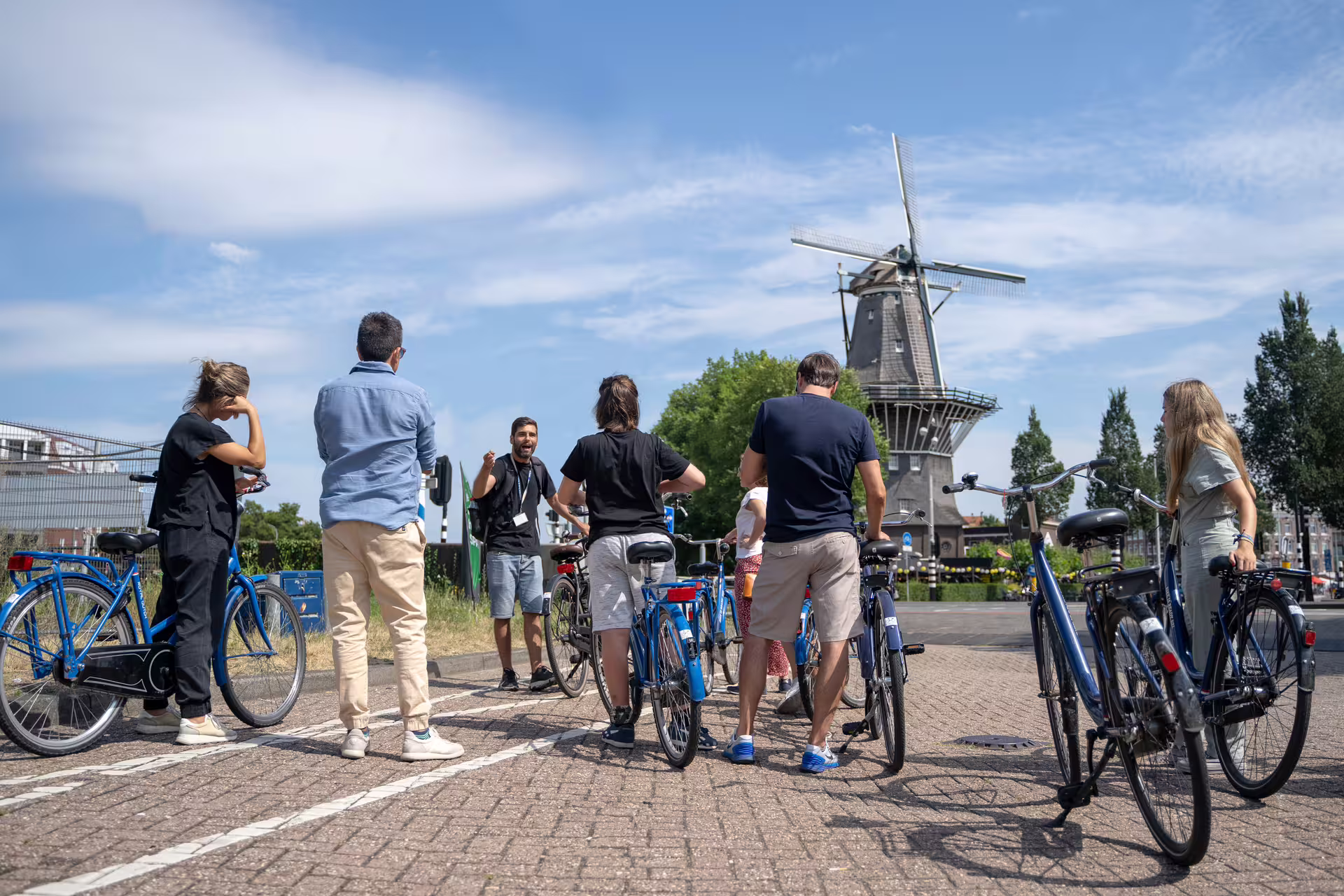 Group of tourists on bikes with guide near iconic windmill during Amsterdam countryside tour, clear blue sky overhead.