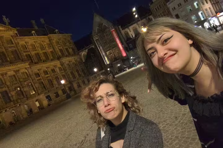 Two women taking a selfie at night in Amsterdam's Dam Square with historic illuminated buildings behind them