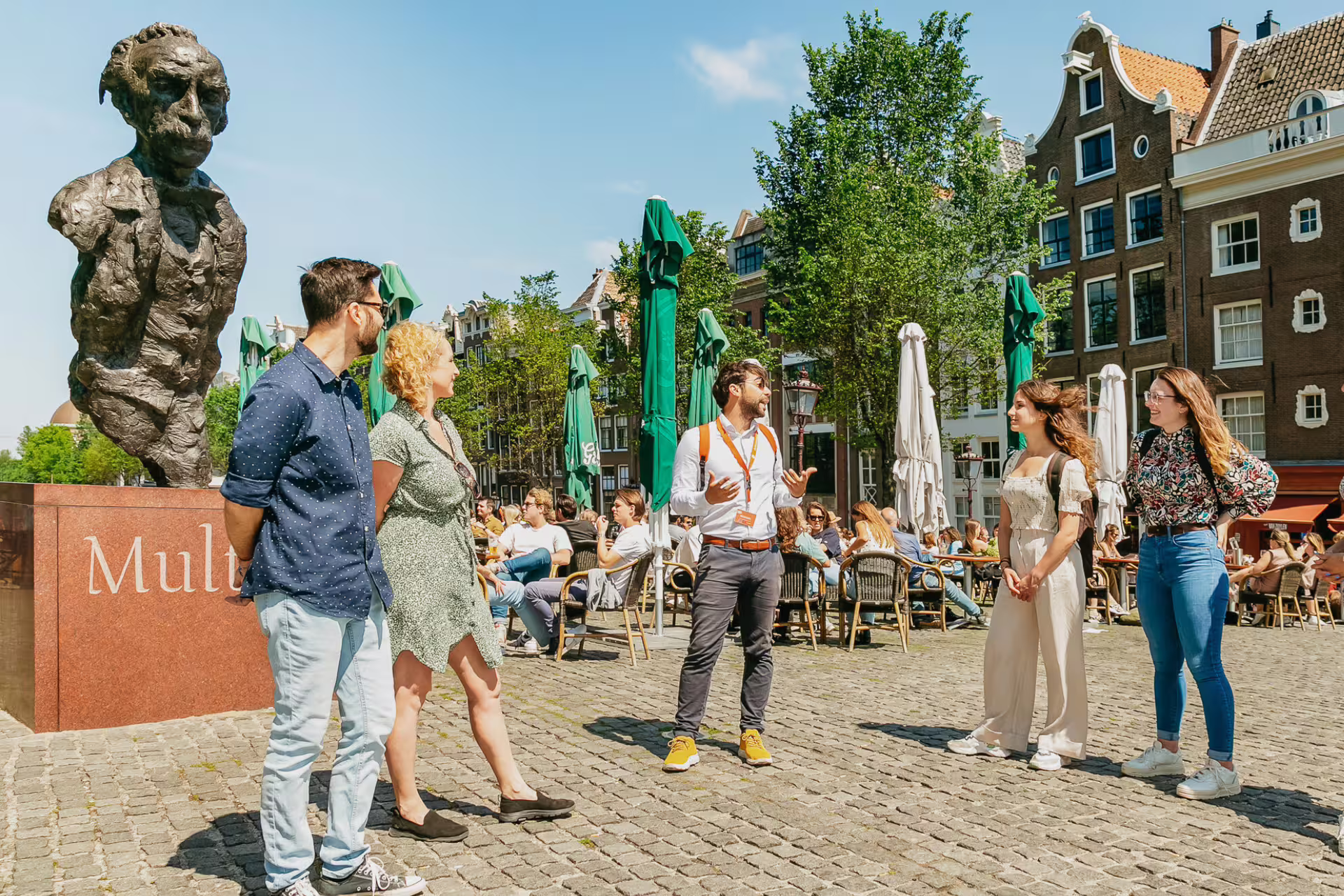 Tour group explores historical Amsterdam with guide near iconic statue in vibrant city square under clear blue skies.
