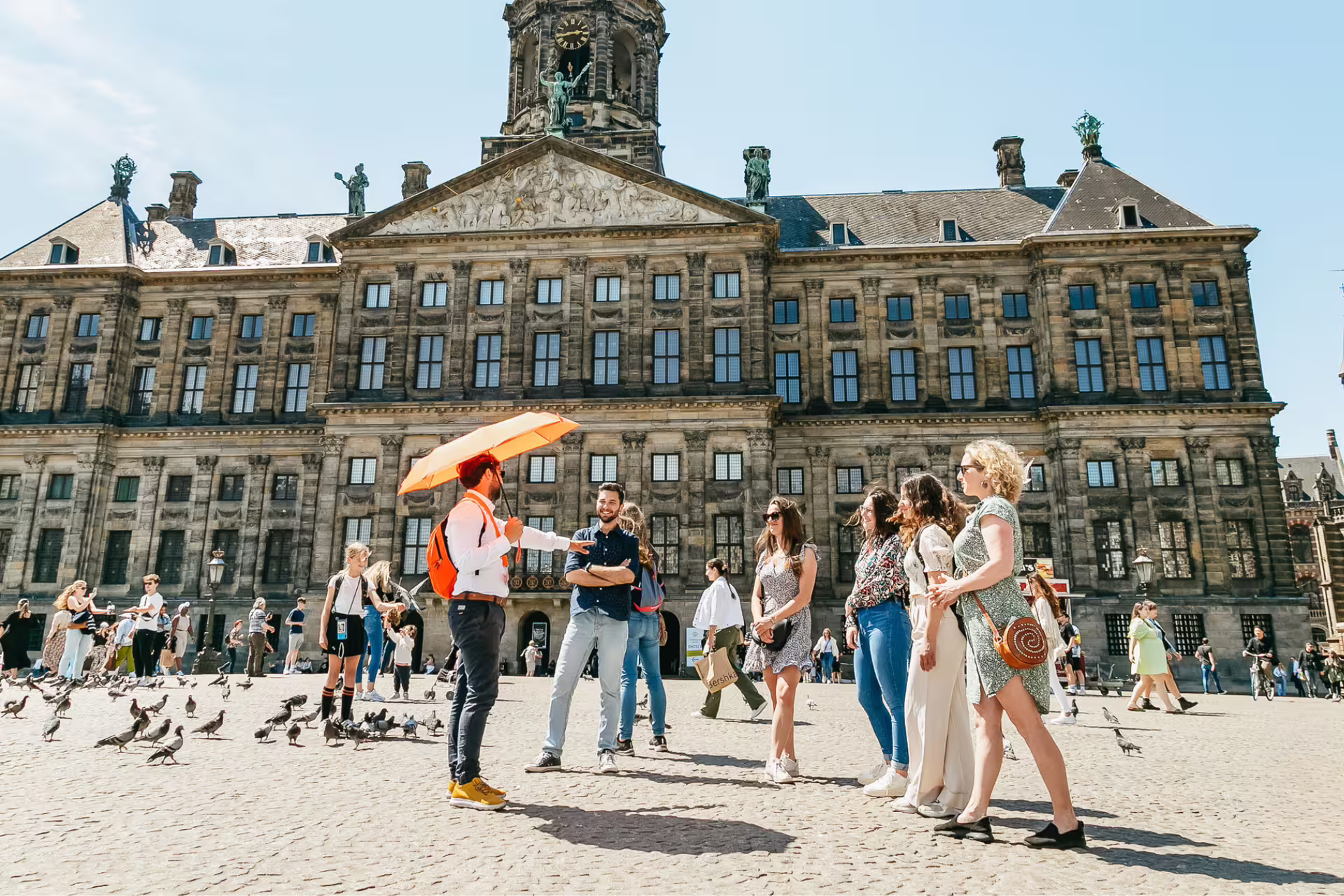 Tour group exploring historical Amsterdam guided by a host with an orange umbrella in front of the Royal Palace on Dam Square.
