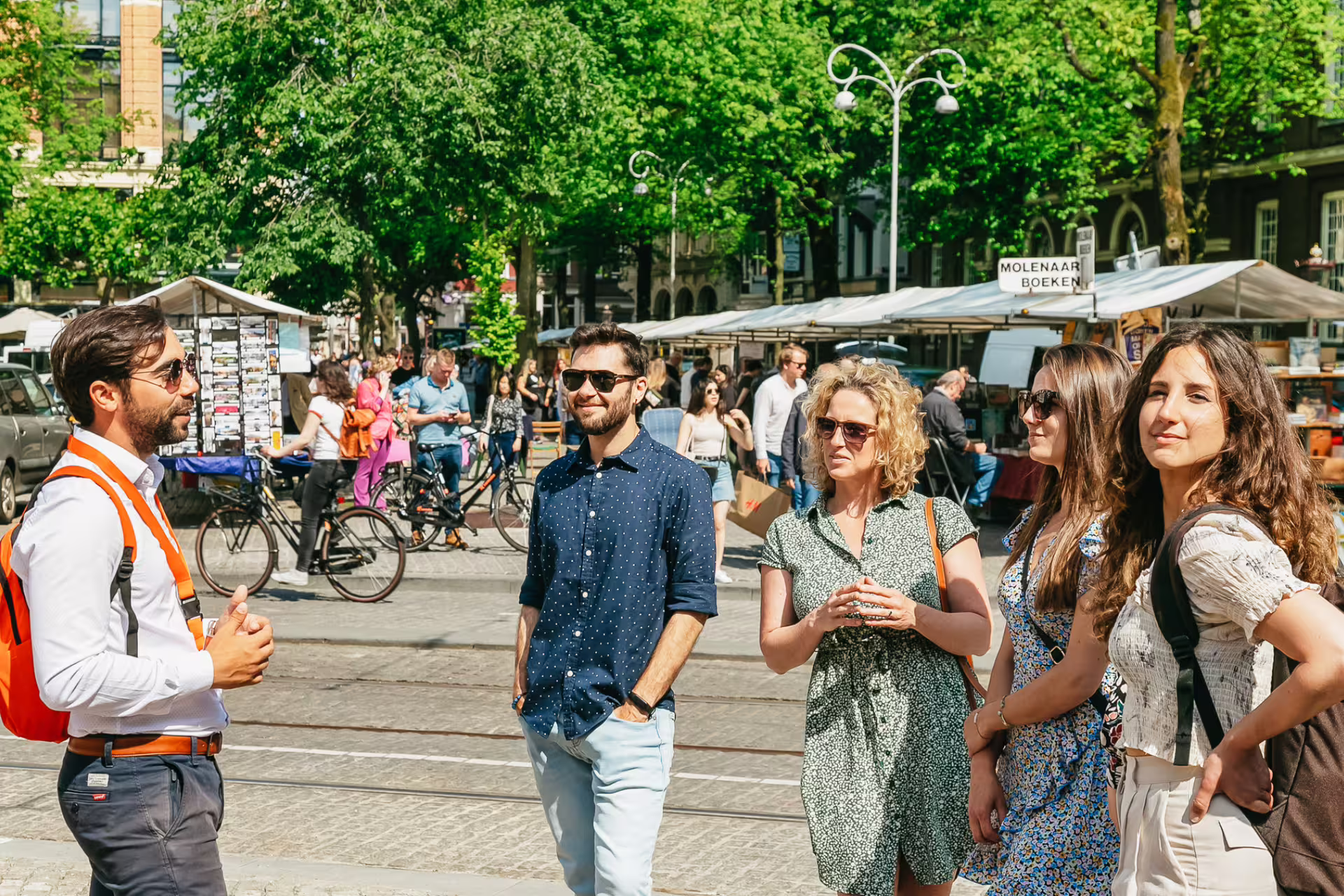 Tour group explores historical Amsterdam market with guide, surrounded by vibrant stalls and bicycles on a sunny day.