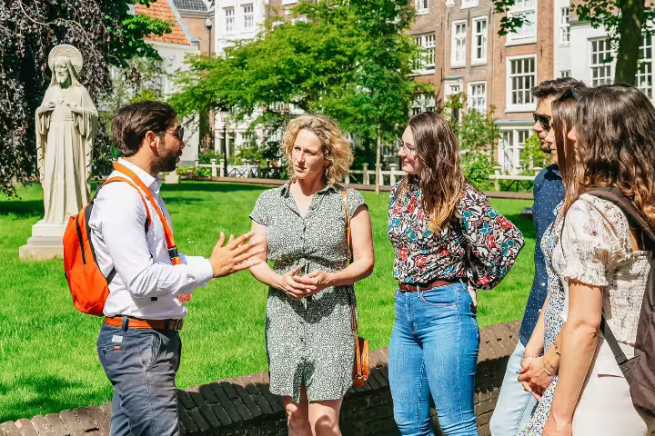 Tour guide discusses historical Amsterdam with visitors in a lush garden near a statue, enhancing cultural exploration.
