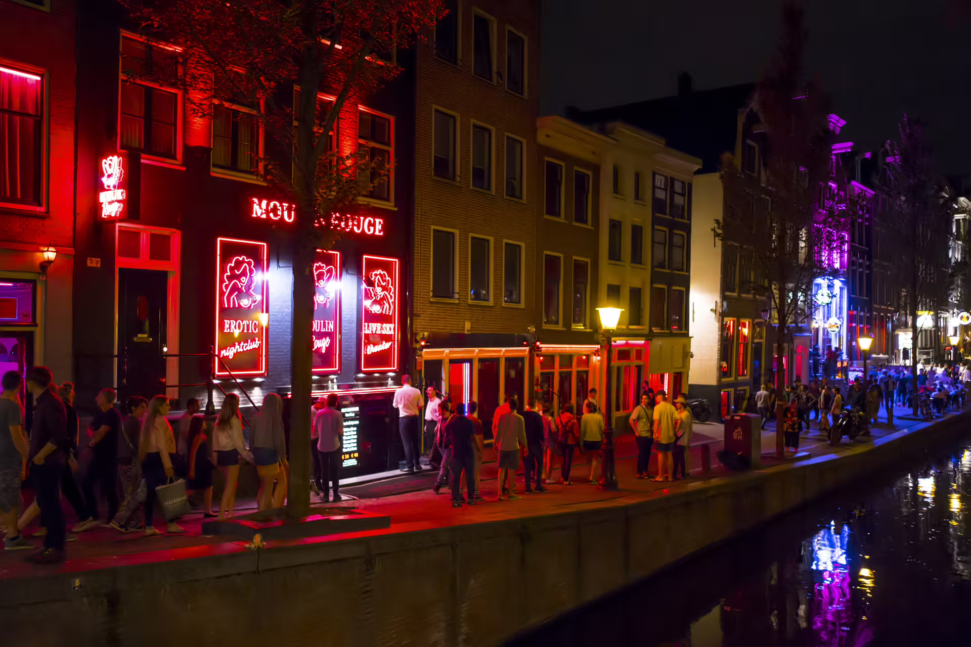 Night view of Amsterdam Red Light District canal with neon-lit windows and crowds, self-guided walking tour with audioguide