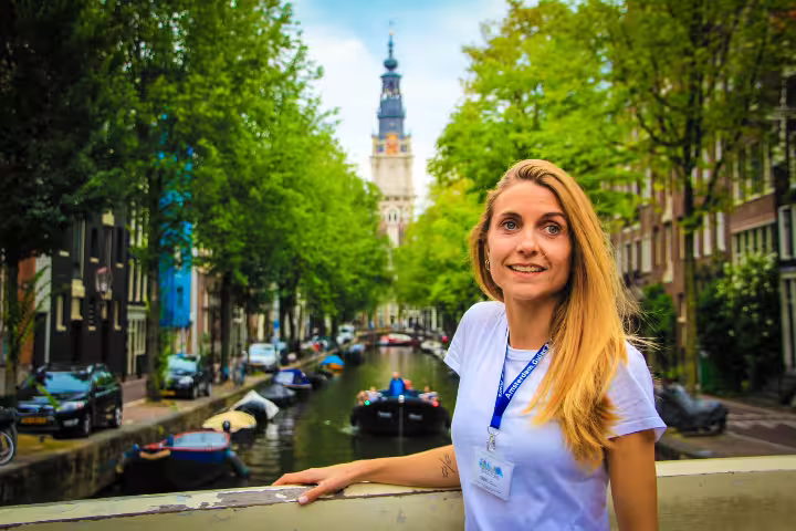 Smiling tour guide on an Amsterdam bridge with scenic canal view, boats, and historic architecture in the background.