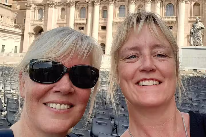 Two smiling women taking a selfie in St. Peter's Square Rome with rows of chairs and basilica behind them