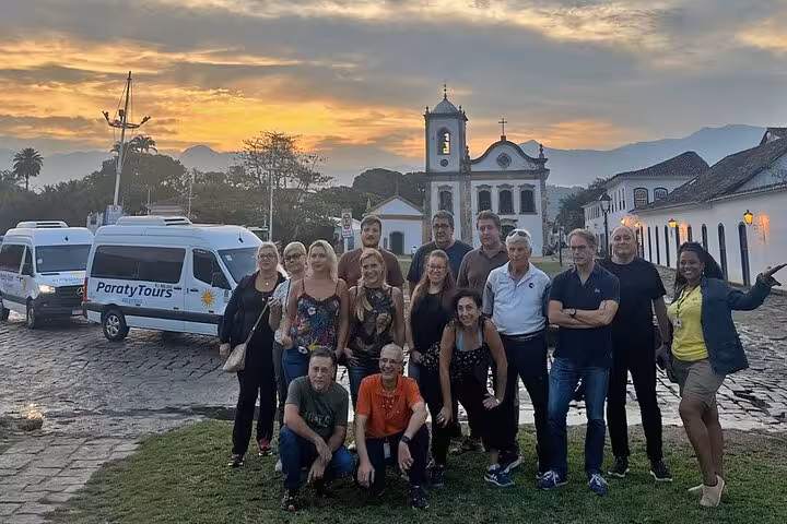 Group of travelers posing by Paraty Tours shuttle with historic church and sunset in Angra dos Reis background.