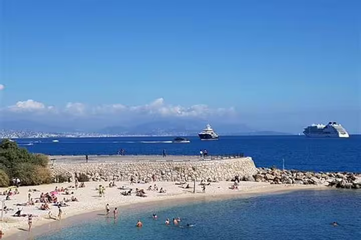 Sunny beach near Antibes with swimmers, stone seawall, and cruise ships on the blue Mediterranean Sea