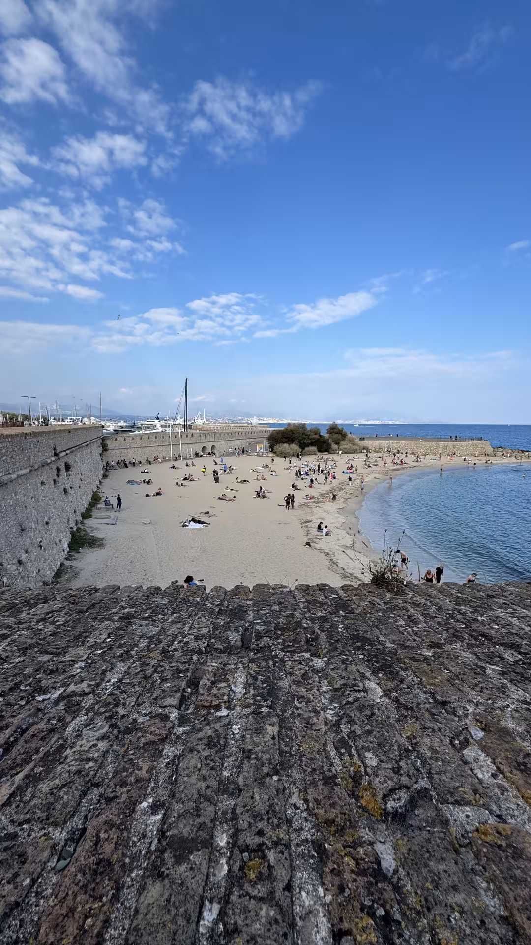 Sandy beach beside ancient stone fortress walls in Antibes, French Riviera, with sunbathers and blue Mediterranean Sea