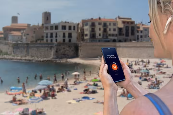Woman holding smartphone with GPS audio guide app at Plage de la Gravette beach in Antibes, France