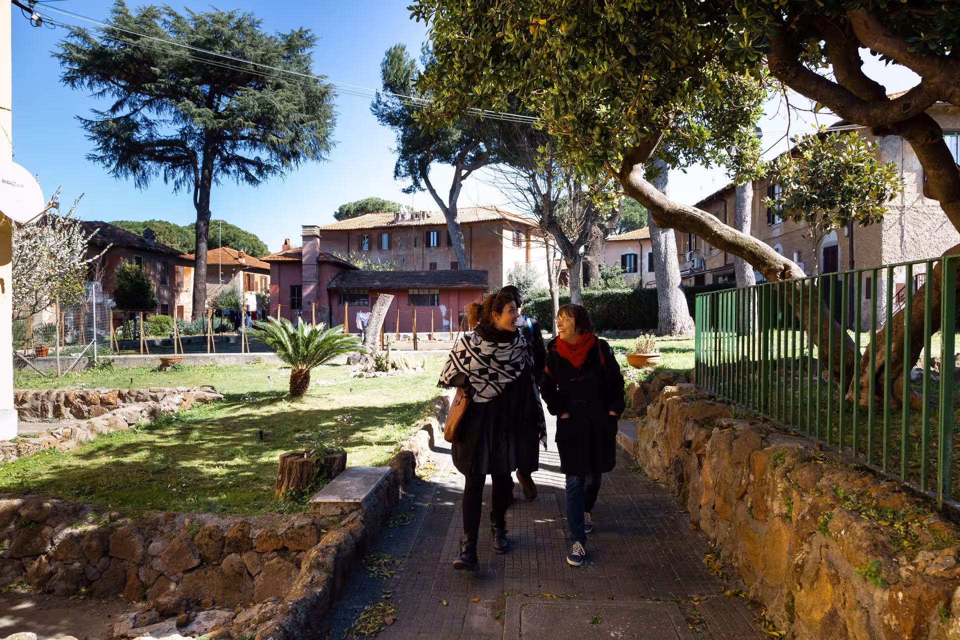 Two women enjoying a scenic walk through Caffarella Valley, surrounded by vibrant greenery and charming homes.