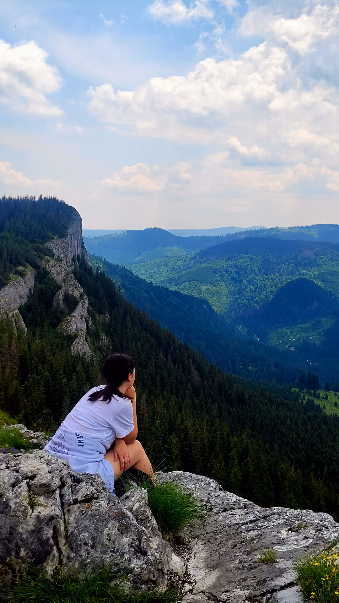 Hiker sitting on rocky cliff overlooking lush green forested valley and mountain peaks in Apuseni