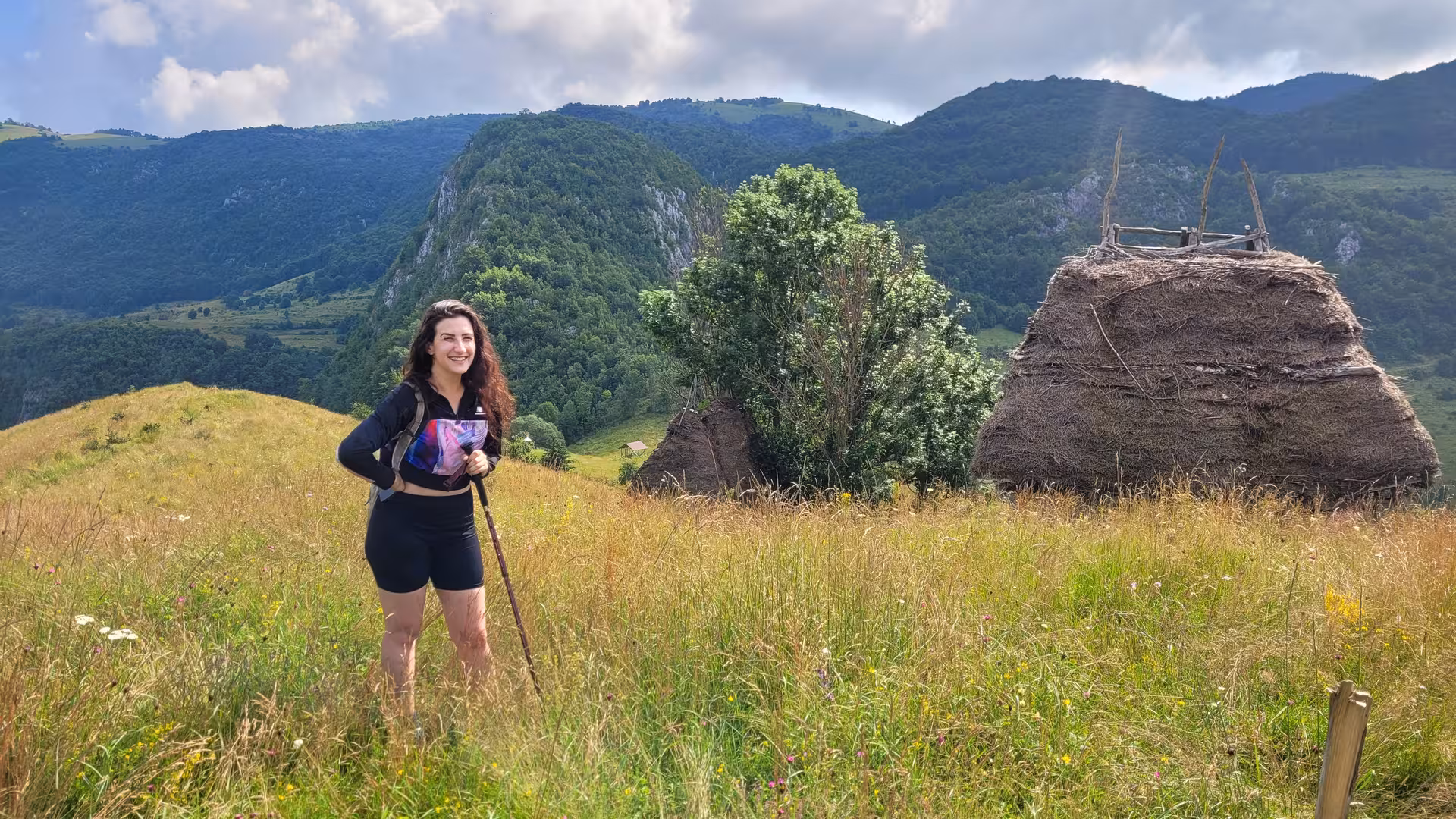 Hiker with trekking pole stands on alpine meadow with haystacks and Apuseni Mountains in background