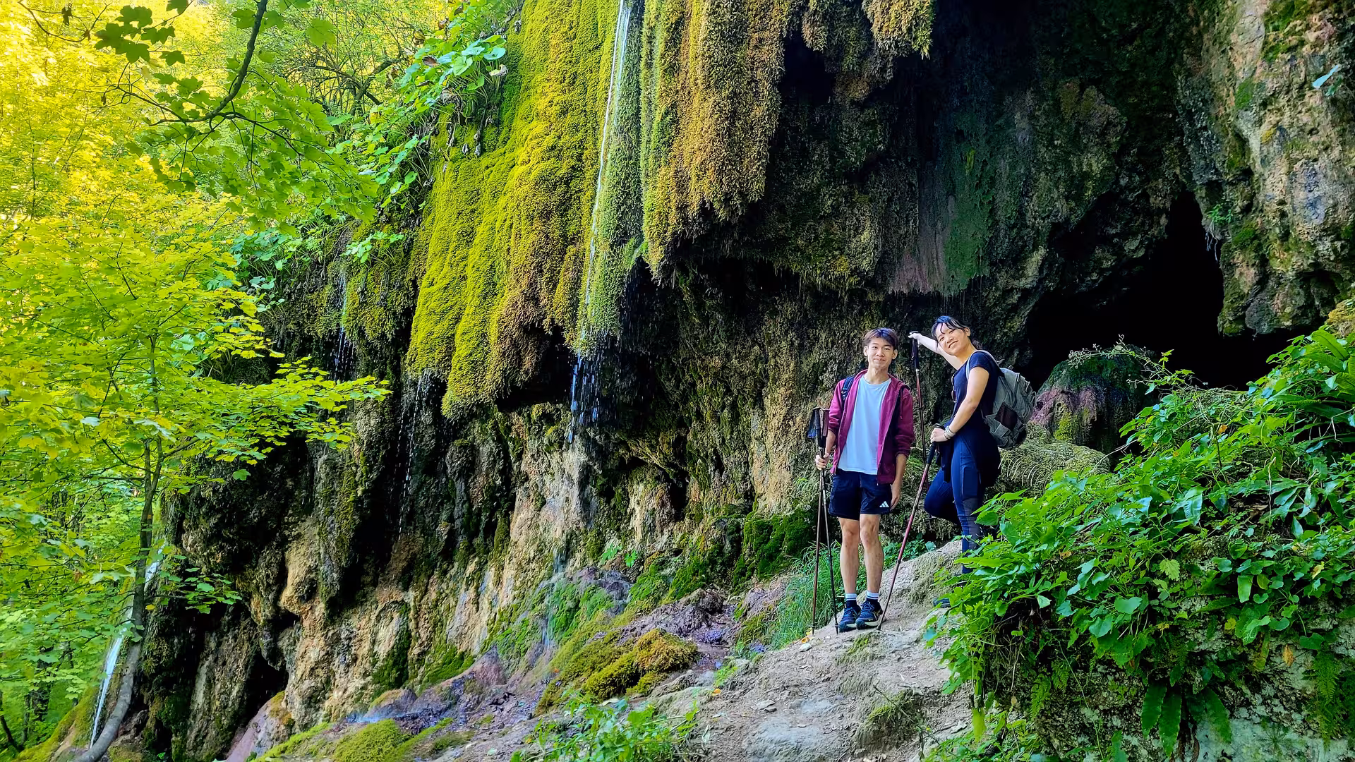Two hikers with trekking poles on mossy cliff trail near waterfall in Apuseni Mountains