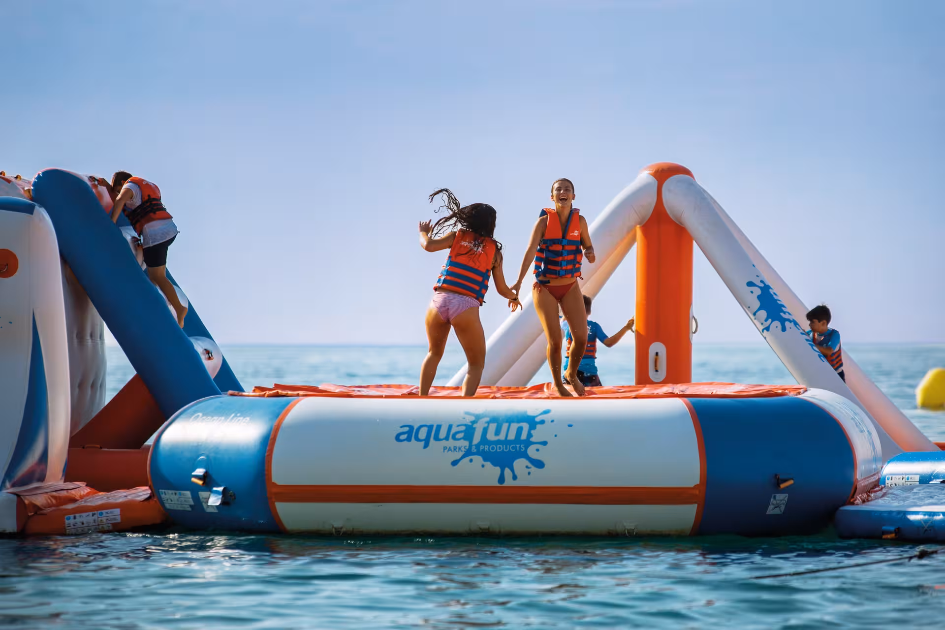 Kids jump and play on an Aquafun inflatable waterpark obstacle course floating on the sea under a clear blue sky.