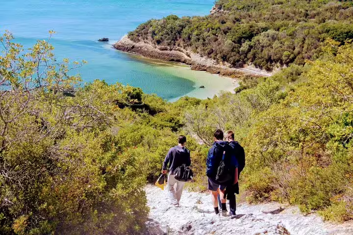 Tourists walking down a scenic trail towards the turquoise waters of Arrábida, ideal for snorkeling and enjoying local wine.