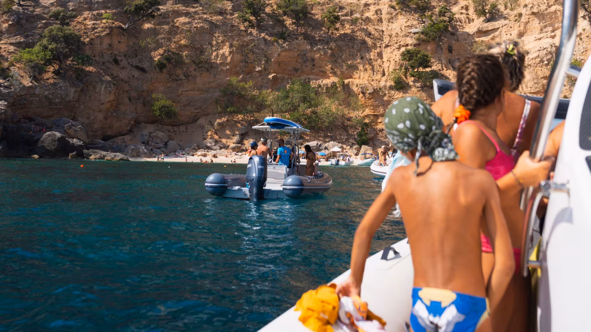 Family enjoying Arbatax RIB trip, approaching Cala Luna's pristine beach surrounded by rocky cliffs.