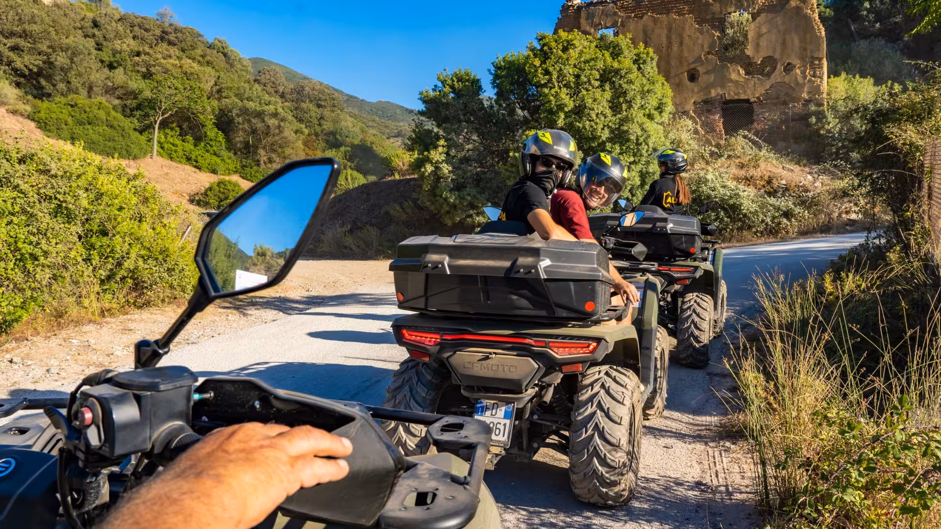 Group of adventurers on ATVs exploring Arbus' scenic trails with lush greenery and historic ruins in the background.